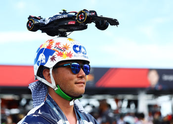 Person wearing a colorful helmet and sunglasses with a Formula 1 car on their head, set against a blurred race track background.